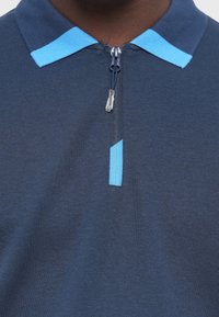 Marineblå polo shirt med lynlås, prydet med lyseblå detaljer på kraven og lynlås-linjen. Det strukturerede stof tilføjer et subtilt mønster.