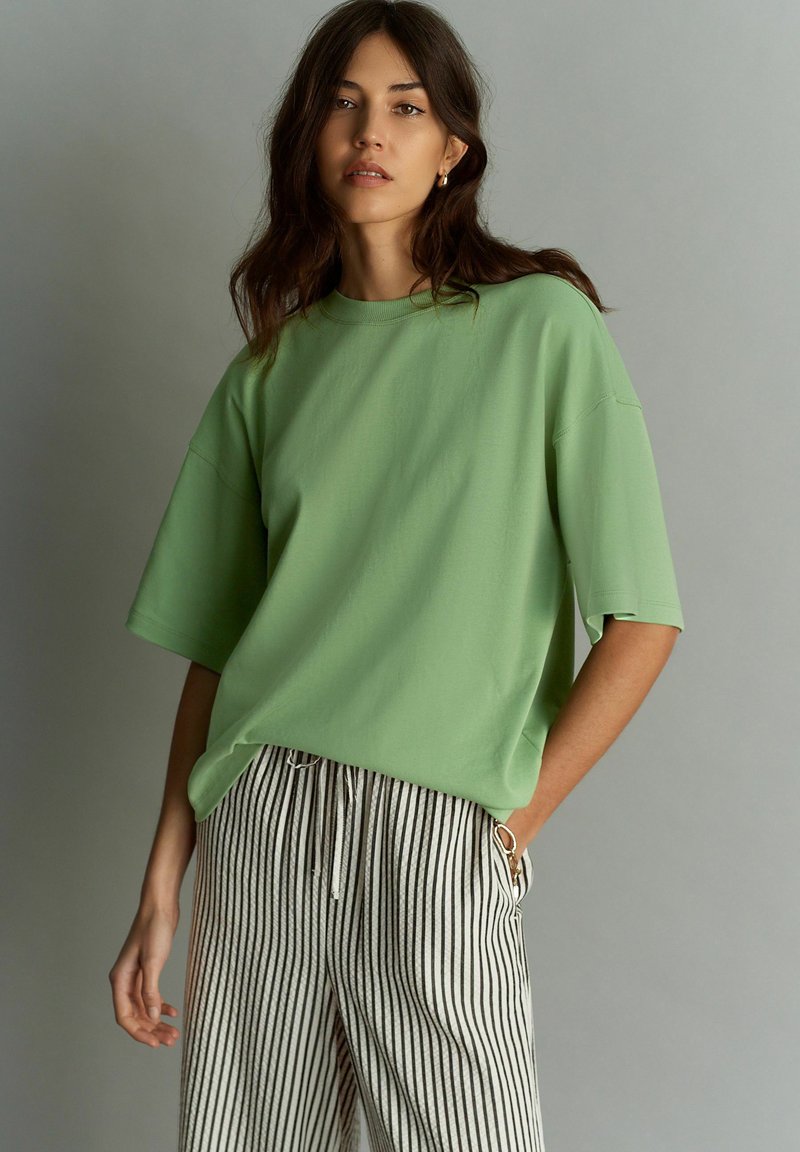 Camiseta oversized verde claro con mangas cortas, combinada con pantalones a rayas en blanco y negro. Textura suave y ajuste relajado.
