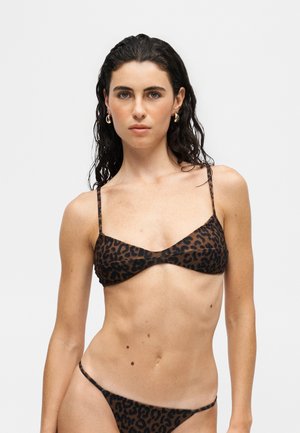 Žena s tmavými mokrými vlasmi, ktorá má na sebe zlaté kruhové náušnice a hnedé bikiny s leopardím vzorom na jednoduchom bielom pozadí.