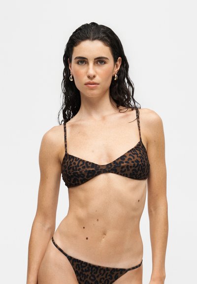Femme aux cheveux foncés mouillés, portant des boucles d’oreilles créoles dorées et un bikini marron imprimé léopard sur un fond blanc uni.
