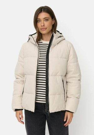 Frau trägt eine hellbeige Steppjacke mit Kapuze, schwarzen Reißverschlussdetails, ein schwarz-weiß gestreiftes Hemd und dunkle Jeans vor weißem Hintergrund.