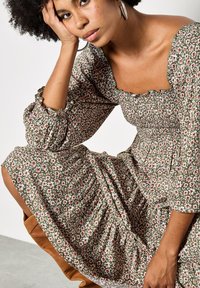 Grünes Blumenmuster-Kleid mit Puffärmeln, ausgestattet mit einem quadratischen Ausschnitt und Rüschenbesatz. Das Material hat eine weiche, geraffte Textur mit kleinen weißen und rosa Blumen.