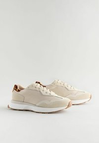 Beige tenisky s texturovaným textilním svrškem, bílou podrážkou a hnědými skvrnitými akcenty na patě. Mají tradiční šněrování a zaoblenou špičku.