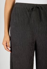 Zwarte geplooide broek met verticale ribbelstructuur, elastische tailleband en koordsluiting. Soepele stof met gedefinieerde patronen en een relaxte pasvorm.