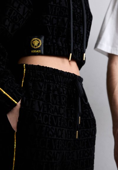 Melnās topiņš un sporta bikses, kas izgatavotas no teksturēta auduma, ar izceltu "VERSACE" logo un zelta akcentiem uz auklām un šuvēm.