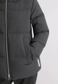 Padded, ciemnoszara kurtka z zapięciem na zamek, kieszeniami po bokach i metką z logo na dolnym brzegu. Materiał ma teksturowany wygląd.