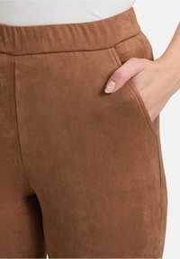 Brązowe zamszowe spodnie o miękkiej fakturze, zwężającym się kroju, elastycznym pasie i bocznych kieszeniach. Detale obejmują przeszywane szwy i gładkie wykończenie.