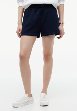 Mujer con pantalones cortos azul marino con cordón, zapatillas blancas y una sudadera blanca, de pie sobre un fondo blanco.