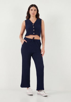 Kvinde står iført navy ærmeløs knap-lukket crop top og matchende vidbenede bukser med hvide sneakers, hænder i lommerne.