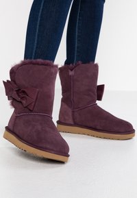 Bottes d'hiver en daim violet avec doublure en fourrure et grands nœuds à l'arrière, portées avec un jean skinny bleu foncé, debout sur un sol blanc lisse.