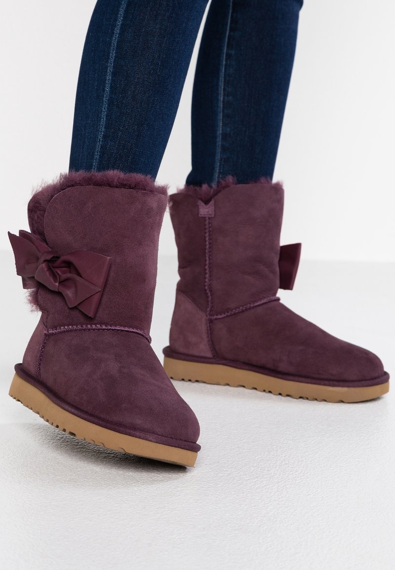 Bottes d'hiver en daim violet avec doublure en fourrure et grands nœuds à l'arrière, portées avec un jean skinny bleu foncé, debout sur un sol blanc lisse.