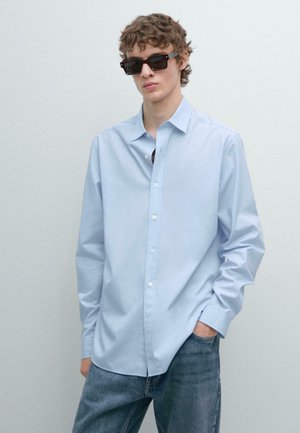 Chemise bleu clair à manches longues avec un col évasé, assortie à un jean foncé et des lunettes de soleil rectangulaires. Texture de tissu lisse.