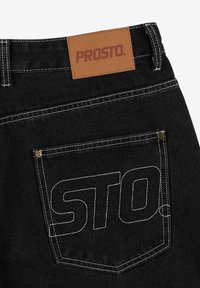 Czarne dżinsy o fasonie denim z jasnobrązową skórzaną łatą w talii. Tylny kieszeń ma białe przeszycia oraz wytłoczone na biało litery "STO".