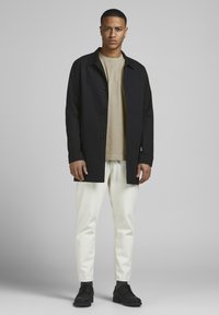 Black cotton jacket with button closure, beige t-shirt underneath, white pants, and black shoes. Smooth texture, minimal design.