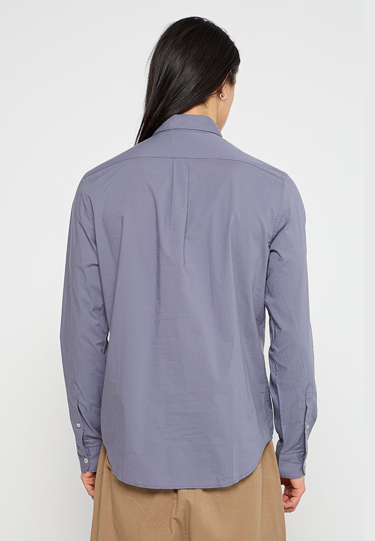 Camisa de manga larga gris con cuello tipo polo, silueta ajustada y puños abotonados. Presenta un pliegue en la parte posterior central para mayor libertad de movimiento.