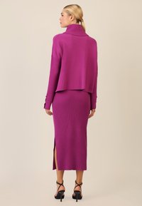 Magenta angeschnittener Pullover mit hohem Kragen, kombiniert mit einem gerippten Midaxi-Rock mit einem seitlichen Schlitz und schwarzen Riemchenabsätzen.