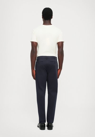 Navy trousers with a slim fit design, featuring back pockets and a smooth texture, paired with a light-colored short-sleeve shirt.