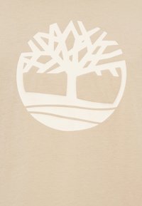 Μπεζ μπλουζάκι με μεγάλο λευκό λογότυπο δέντρου με γεωμετρικά κλαδιά και καμπυλωτές γραμμές εδάφους μέσα σε κυκλικό περίγραμμα. Απαλή υφή.