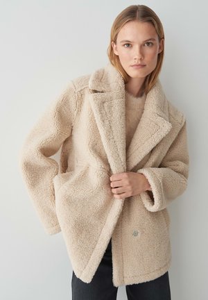 Beige fleecejas met een sjawlcollar, voorzakken en een iets oversized pasvorm, gedragen over een lichte trui, gepresenteerd tegen een neutrale achtergrond.