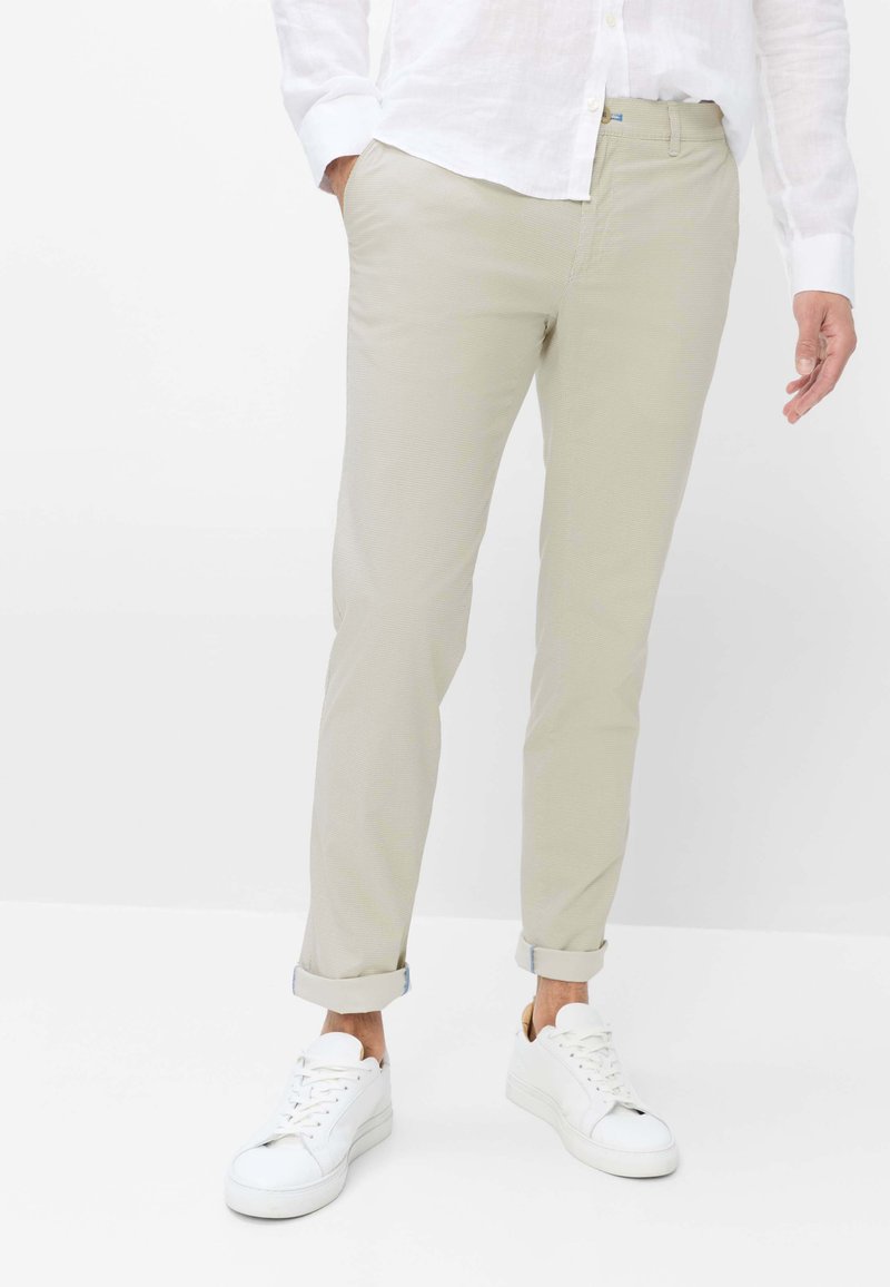 Uomo che indossa pantaloni beige slim fit con risvolti e sneakers bianche, mostrato dalla vita ai piedi su sfondo bianco.
