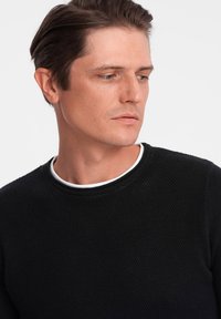 Black textured sweater with a round neckline, featuring a white inner collar. Close-up view focusing on fabric and detailing.