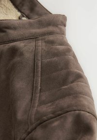 Tela de ante marrón con un patrón acolchado y textura suave. Presenta un cuello alto y costuras limpias a lo largo de las costuras.