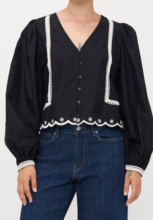 Czarna bluzka na guziki z bufkami na długich rękawach, z białą koronkową lamówką na krawędziach i mankietach, w połączeniu z niebieskimi jeansami.