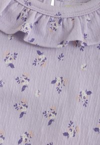 Lavendelfärgad tyg med texturerad yta, prydd med små blommönster i vitt, lila och persika. Volangdetalj vid halslinningen.