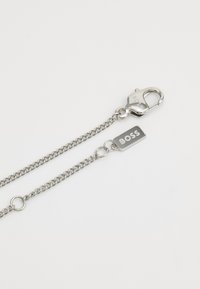 Accessoire en chaîne argentée avec un fermoir en homard. Comprend une étiquette rectangulaire gravée avec "BOSS". Fini métallique élégant avec des maillons entrelacés.