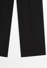 Onderste gedeelte van zwarte, rechte pantalon tegen een witte achtergrond.