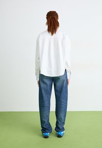Persona con trenzas de cabello de pie de espaldas, vistiendo una camiseta blanca a rayas, jeans azul holgados y zapatillas azul y blanca sobre un suelo verde.
