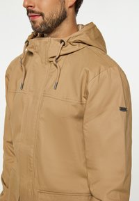 Beige huva jacka med en slät textur, justerbara snoddar och en dragkedjedetalj på ärmen. Framfickor synliga.