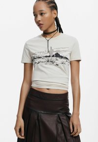 Béžové bavlnené tričko s grafickým zobrazením čiernej púštnej krajiny, doplnené tmavou koženou plisovanou sukňou. Dizajn obsahuje textový detail.