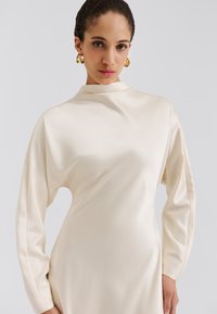 Vestido color crema de satén con cuello alto y mangas largas y suavemente drapeadas. Presenta una textura suave y un diseño elegante y minimalista.