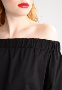 Top nero scollato con spalle scoperte, con scollatura elastica arricciata, texture liscia e tessuto drappeggiato, che mette in evidenza un fit rilassato.