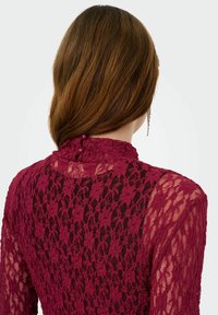 Blouse en dentelle marron avec col montant à volants, manches longues transparentes et boutons décoratifs au dos. Motif floral texturé sur toute la surface.