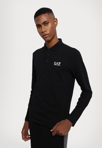 Polo a maniche lunghe nera con colletto abbottonato e logo EA7 bianco. Tessuto liscio, design aderente e senza motivi o dettagli visibili.