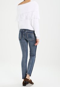 Biała sweter bez ramiączek z frędzlami, zestawiony z niebieskimi skinny jeansami z ciemnym paskiem po boku oraz brązowymi butami w panterkę.
