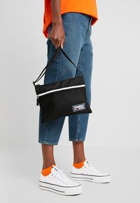 Bustina nera con zip, caratterizzata da un design a rete, con una striscia bianca e il logo Tommy Jeans. La persona indossa jeans blu e scarpe bianche con accenti arancioni.