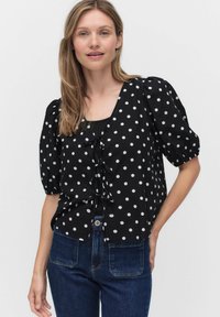 Svart kortärmad blus med vita prickar och V-ringning, med ett framknutet detaljer och avslappnad passform. Parat med blå jeans.