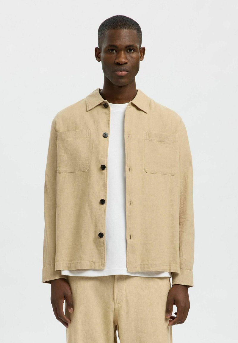 Giacca leggera beige con colletto, due tasche sul petto e chiusura con bottoni. Indossata sopra una t-shirt bianca, presenta una vestibilità comoda.