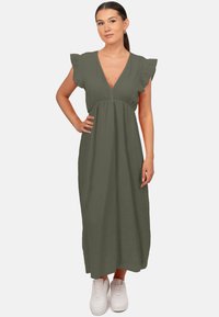 Robe maxi en vert olive avec des manches volantées, décolleté en V, tissu texturé et silhouette fluide, associée à des baskets blanches.