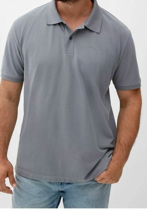 Homme portant un polo gris uni à manches courtes et un jean bleu clair, une main dans la poche sur fond blanc.