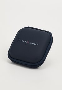 Custodia rigida blu navy con chiusura a zip. Mostra il logo "TOMMY HILFIGER" in bianco e rosso. Texture liscia con bordi arrotondati.