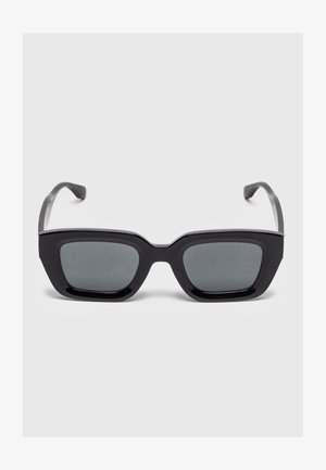 Gafas de sol rectangulares negras con montura gruesa y lentes tintadas oscuras, descansando sobre una superficie gris claro.