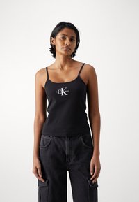 Black tank top with thin straps and a fitted design, featuring a white logo centered below the neckline. Paired with dark cargo pants.