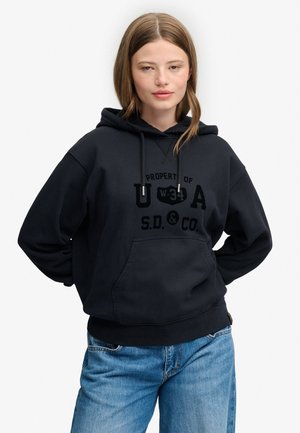 Ung kvinde iført en sort hættetrøje med trykt tekst og blå jeans, stående mod en ensfarvet hvid baggrund.