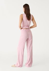 Femme avec de longs cheveux bruns portant un haut floral sans manches rose noué dans le dos et un pantalon large assorti, se tenant avec les mains sur les hanches.