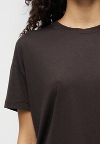 T-shirt à manches courtes marron foncé en tissu doux. Caractérisé par un col rond et une coupe décontractée, avec des détails de couture visibles sur les coutures.