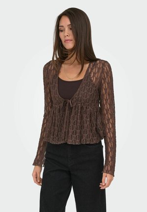 Mujer con blusa marrón de encaje de manga larga sobre un top oscuro y pantalones negros de cintura alta, de pie frente a un fondo liso.